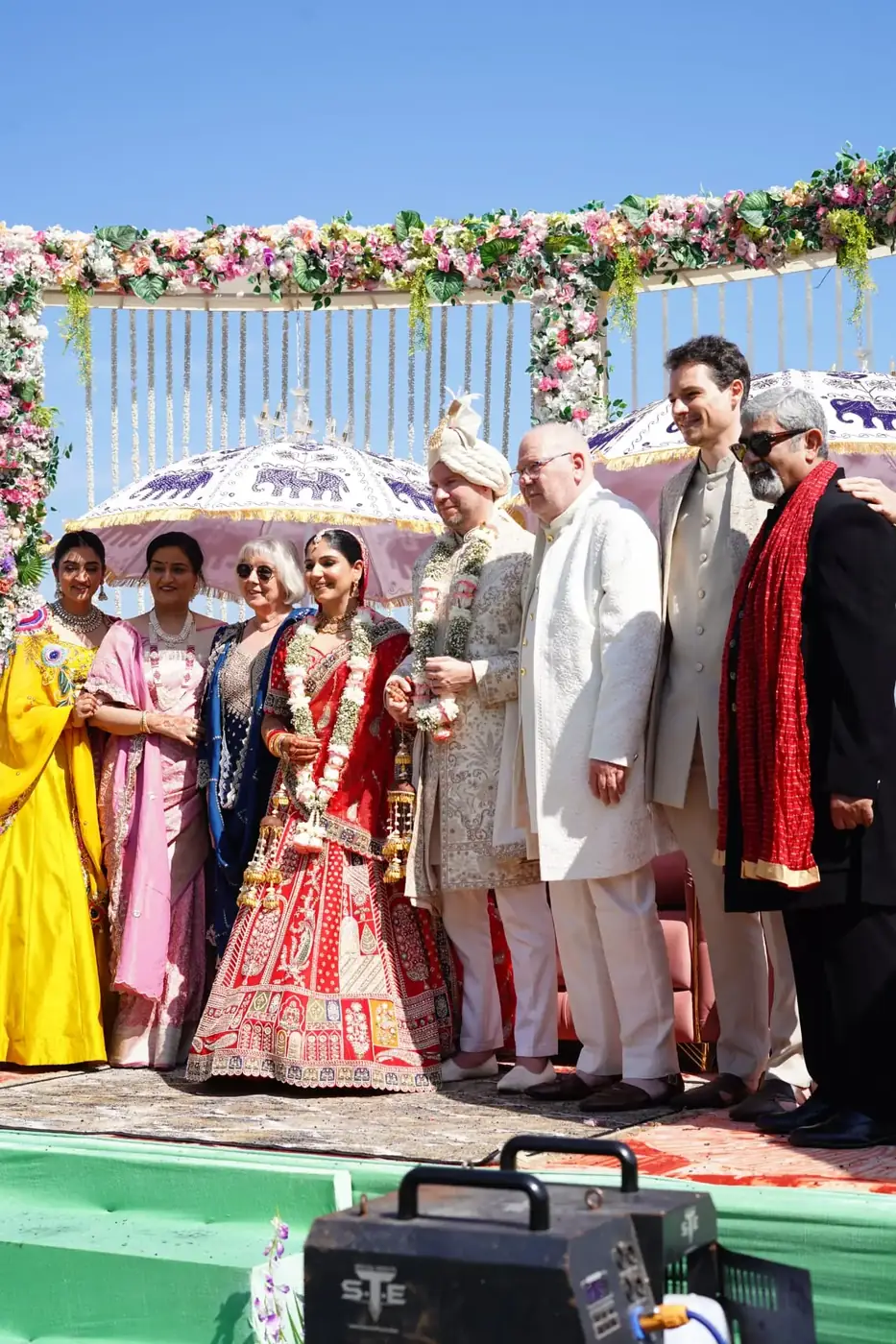 Bride and groom with family beneath decorative parasols at the mandap